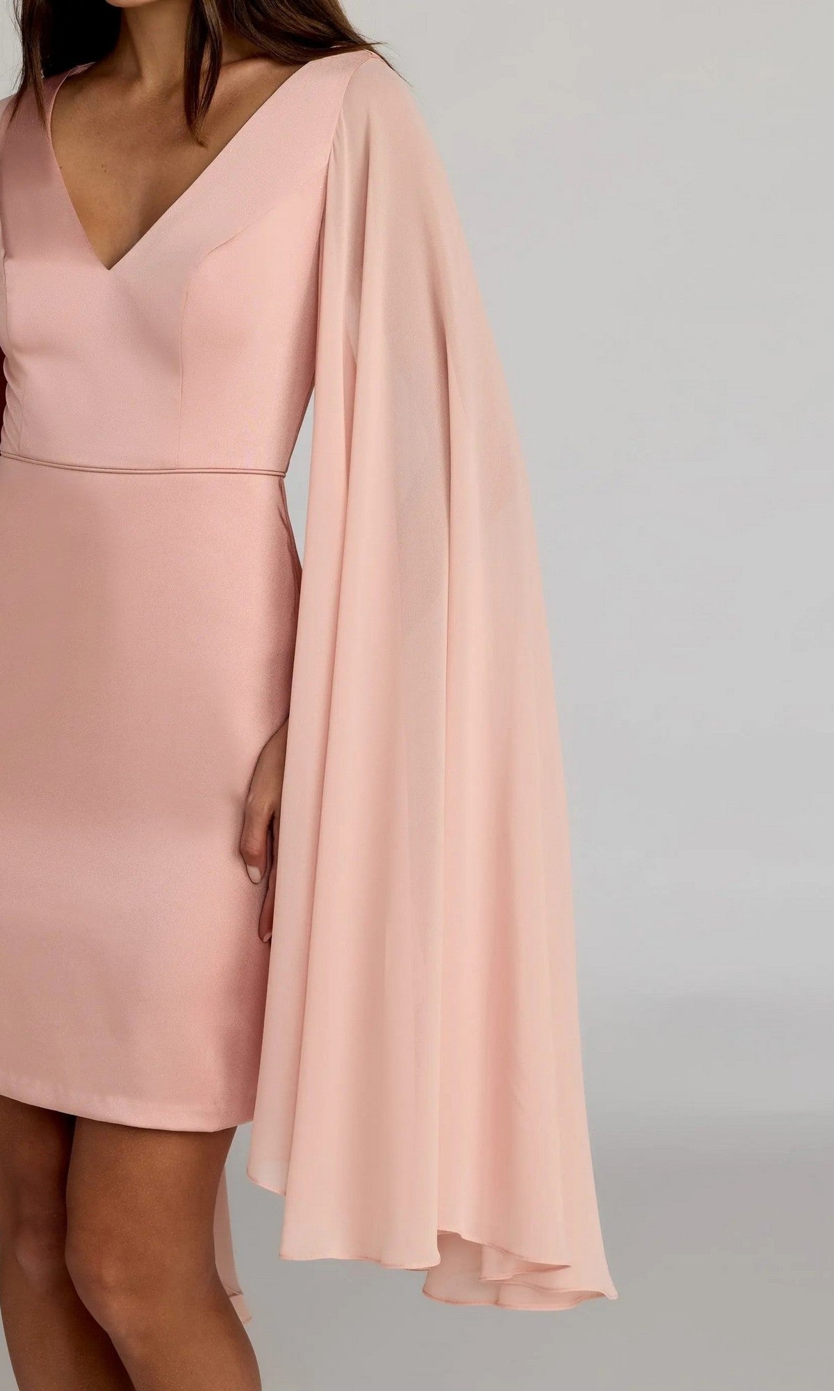 A woman wears the Mac Duggal 55358 short semi-formal dress, featuring light pink V-neck styling with a fitted bodice and one-sided long chiffon cape sleeve. The elegant look stands out against a plain, neutral background.
